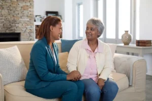 senior woman and younger woman smiling at each other while holding hands on a sofa.