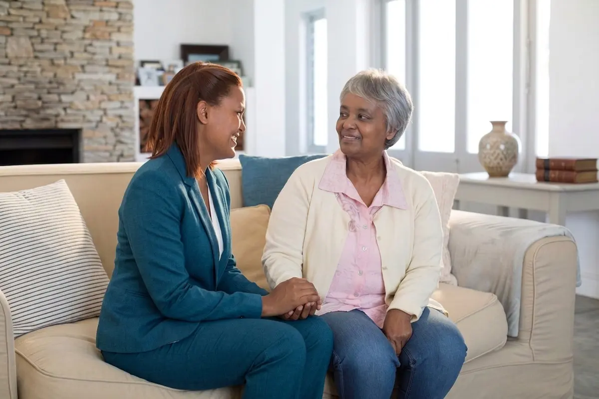 senior woman and younger woman smiling at each other while holding hands on a sofa.