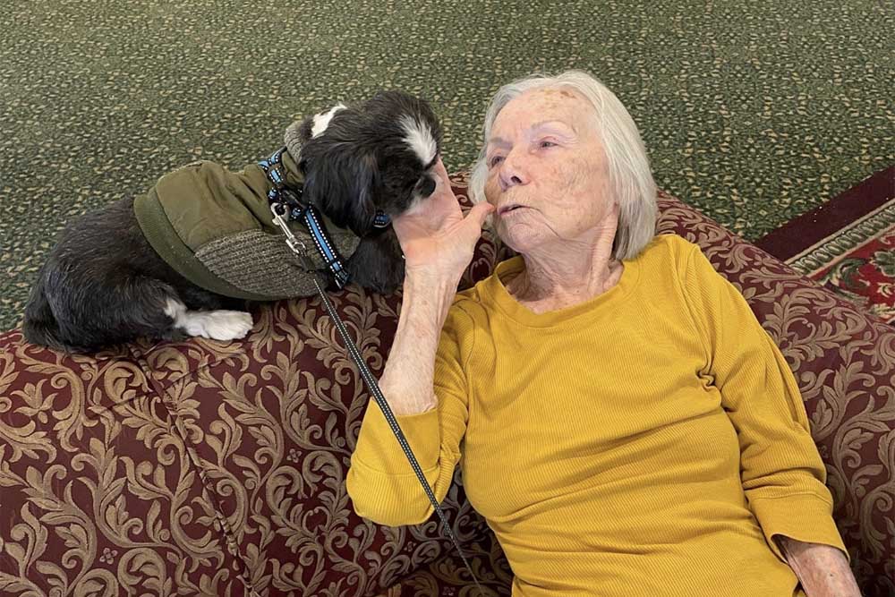 Pegasus Senior Living | Senior woman laying on couch and trying to kiss a small black dog