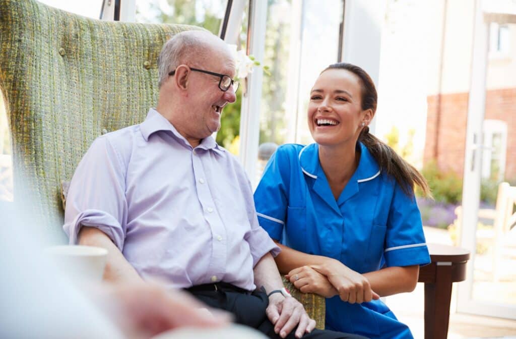 Pegasus Landing of South Plains | An older adult man in a memory care facility sitting on a chair smiling and having a conversation with a nurse.