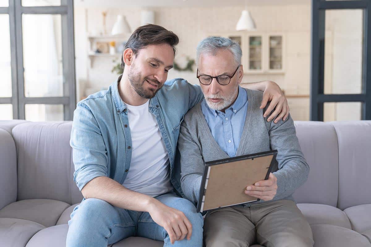 The Farrington at Tanglewood | Caring loving adult son hugging embracing his old elderly senior father while he is showing his family photograph