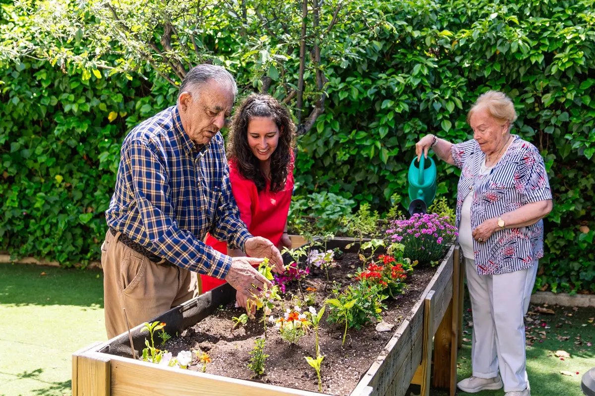 Elderly residents with a caregiver planting and watering flowers in a raised garden bed at a retirement home facility