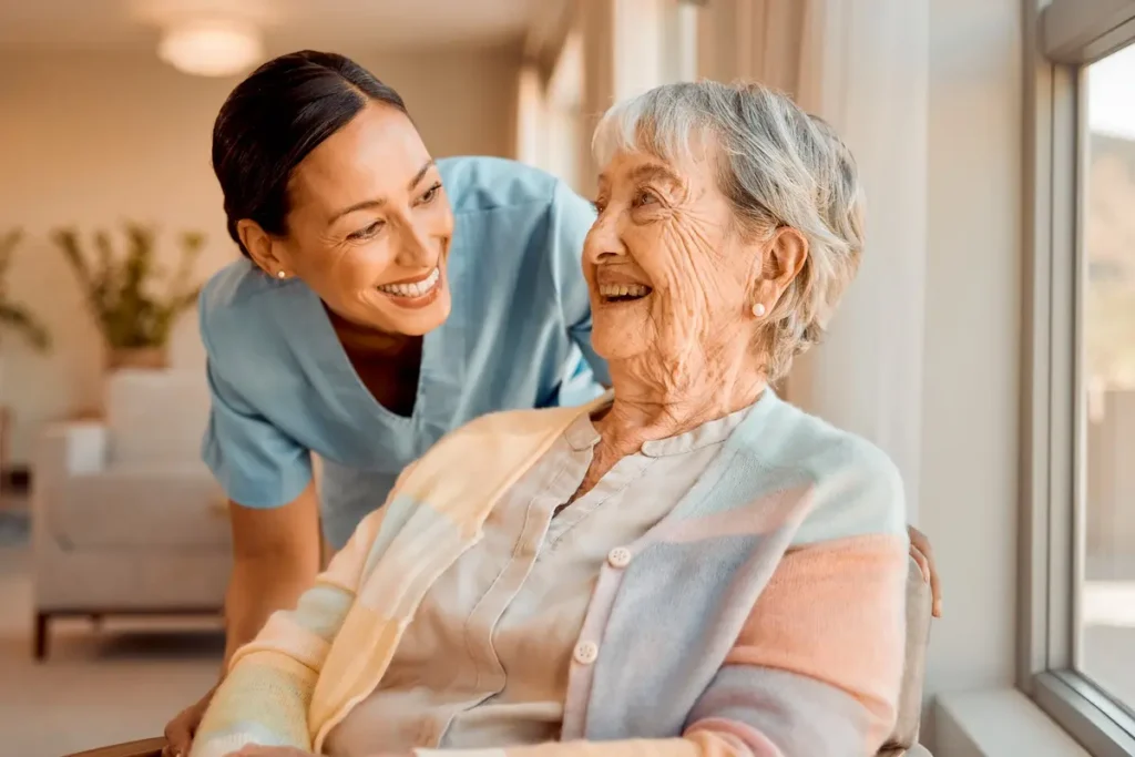 caregiver and seated senior woman smiling at each other beside a sunny window.
