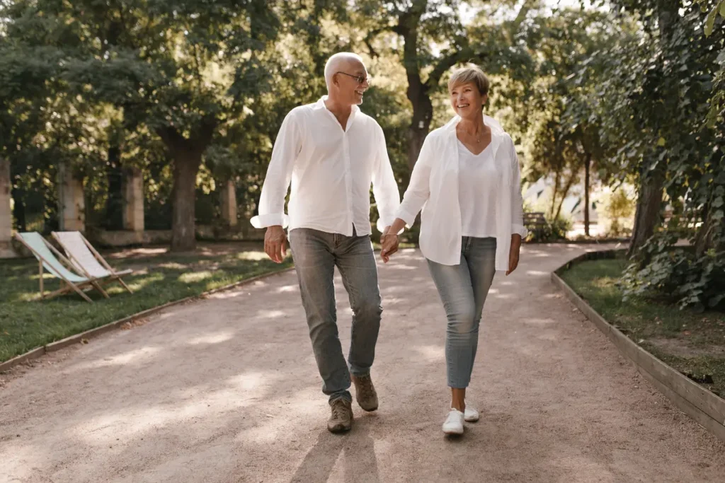 Senior couple smiling and holding hands while walking along a tree-lined park path.