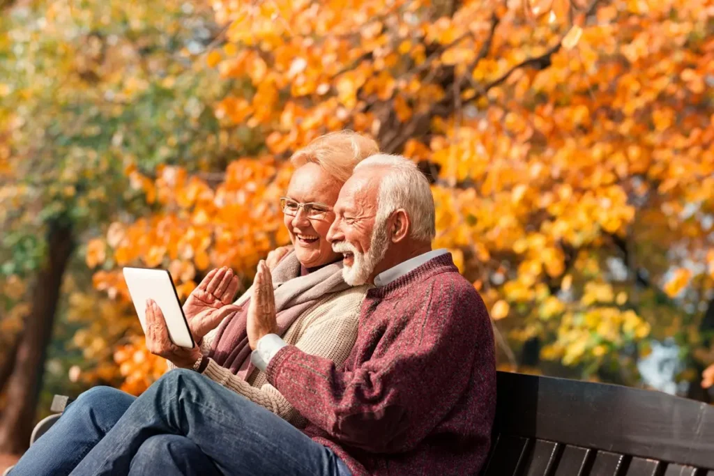Happy senior couple talking, waving, smiling and say Hi their grandkids via tablet in autumn city park